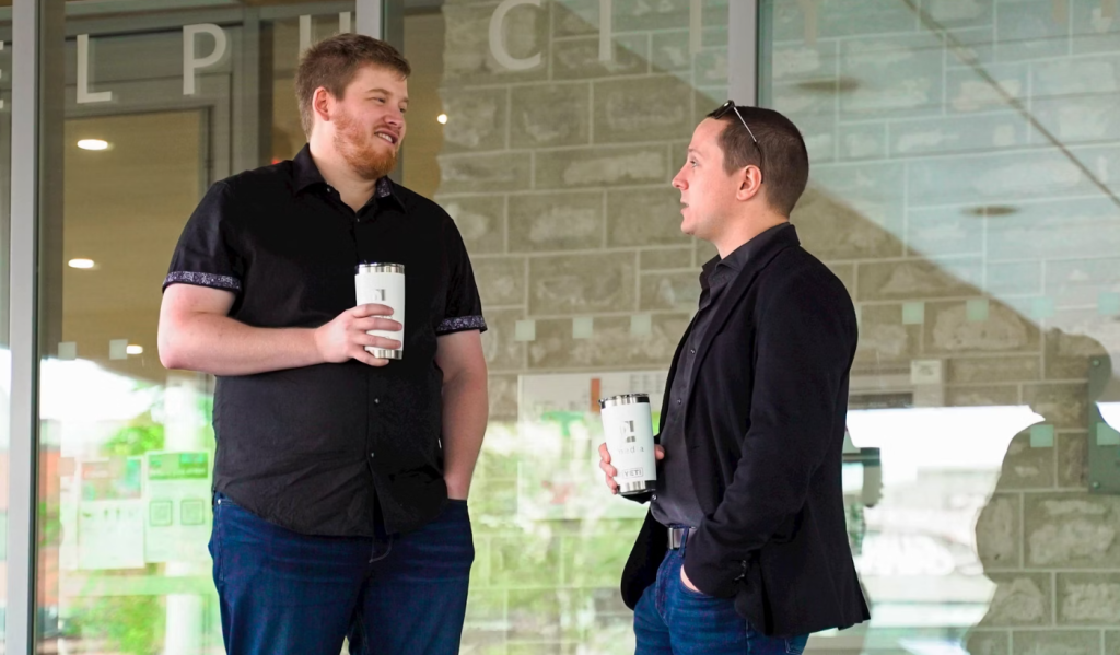 Alt text: "Two men discussing the latest Creator Point podcast while holding branded drinks outside a building with glass doors and a stone facade, both dressed in dark clothing, one wearing sunglasses on his head. Creatorpoint – Creator Knowledge, Simplified. The best resources, podcast summaries, and learning for creators.