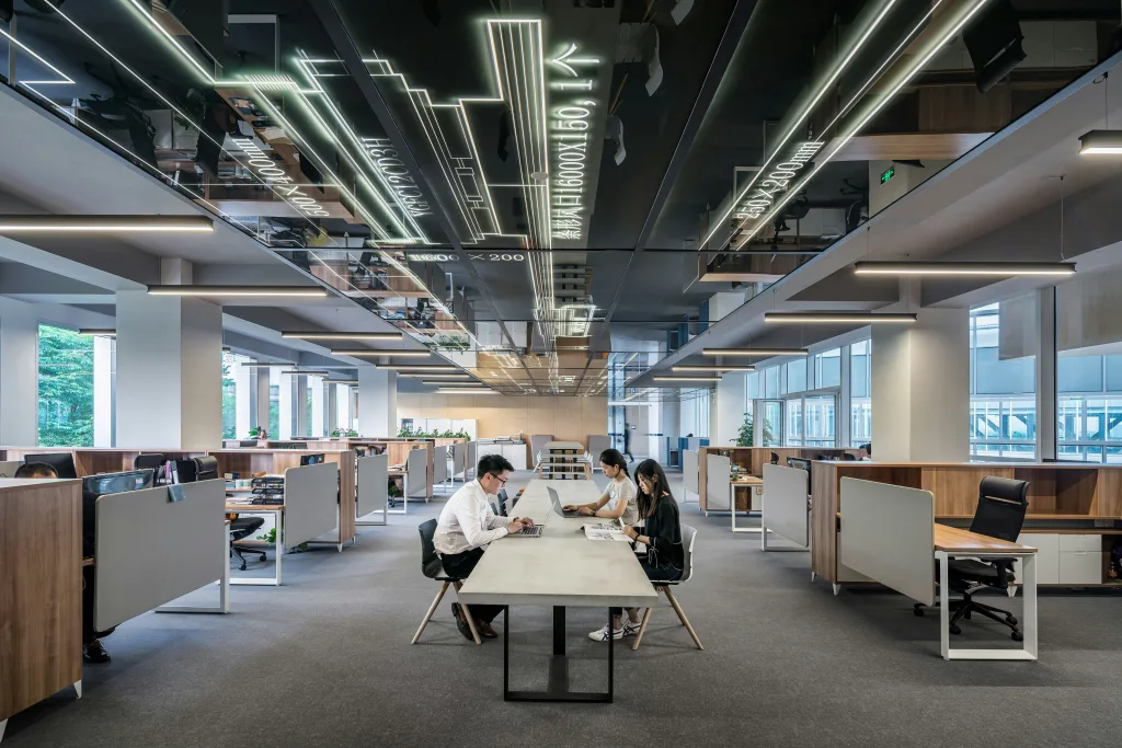 Modern office space with multiple workstations, three colleagues discussing a new podcast summary at central table, reflective ceiling and linear lighting, large windows providing natural light, visible cubicles in background. Creatorpoint – Creator Knowledge, Simplified. The best resources, podcast summaries, and learning for creators.
