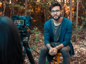 Alt text for SEO optimization: "Content creator in glasses wearing a blue cardigan and green shirt, filming in a forested area for CreatorPoint with camera on tripod among trees. Creatorpoint – Creator Knowledge, Simplified. The best resources, podcast summaries, and learning for creators.