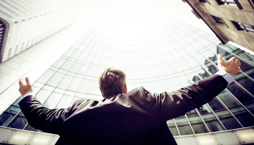 Business professional in suit triumphantly facing modern skyscraper with outstretched arms, symbolizing success and aspiration, viewed from below against a backdrop of bright sky and curved glass facade. Creatorpoint – Creator Knowledge, Simplified. The best resources, podcast summaries, and learning for creators.