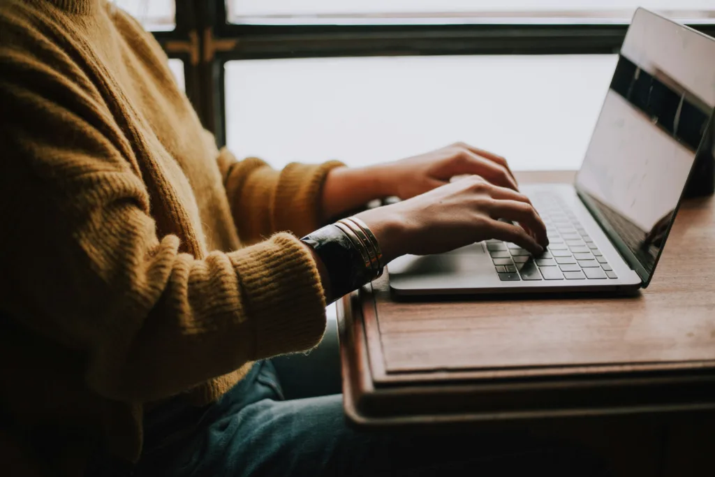 Alt Text: "Person in mustard yellow sweater typing on silver laptop crafting podcast summary at wooden table. Sunlit window with crossbars in background. Dark wristband on left wrist. Working from home, remote work setup, cozy home office. Creatorpoint – Creator Knowledge, Simplified. The best resources, podcast summaries, and learning for creators.