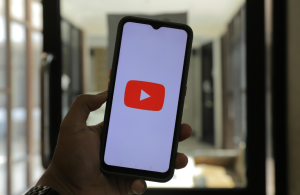 Alt text: "Smartphone displaying YouTube logo on screen, held in a cozy room with blurred windows and furniture background, ideal for watching podcasts or Creator Point content. Creatorpoint – Creator Knowledge, Simplified. The best resources, podcast summaries, and learning for creators.