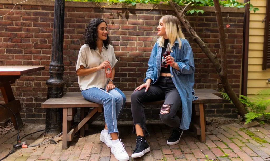 Two women sitting on a wooden bench against a brick wall. One, with curly hair and a beige top, holds a bottle. The other, with straight hair and denim jacket, holds a can. They discuss writing tips for beginners outdoors surrounded by greenery. Creatorpoint – Creator Knowledge, Simplified. The best resources, podcast summaries, and learning for creators.
