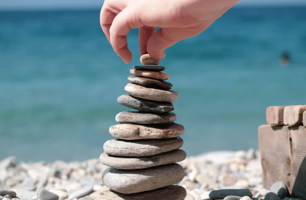 A hand adjusts the top stone of a balanced stack on a pebbled beach with calm blue sea and clear skies in the background, perfect for inspiring beginner writing tips. Creatorpoint – Creator Knowledge, Simplified. The best resources, podcast summaries, and learning for creators.