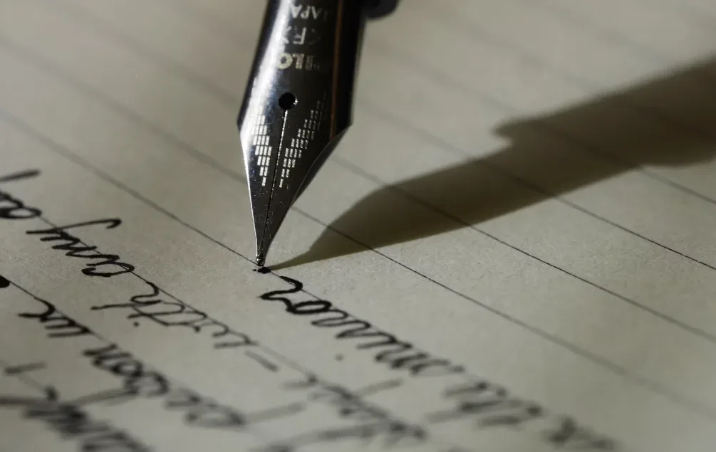 Close-up of a fountain pen nib writing on lined paper, with a shadow cast by the nib and cursive handwritten text partially blurred. Ideal for those searching for writing tips for beginners, this image captures the essence of starting mid-sentence. Creatorpoint – Creator Knowledge, Simplified. The best resources, podcast summaries, and learning for creators.