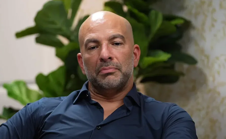 Bald man with beard in navy blue shirt, sitting in front of green leafy plants, looking straight ahead with a neutral expression, contemplating writing tips for beginners. Ideal setting for fresh writing ideas and creative inspiration among natural greenery. Creatorpoint – Creator Knowledge, Simplified. The best resources, podcast summaries, and learning for creators.