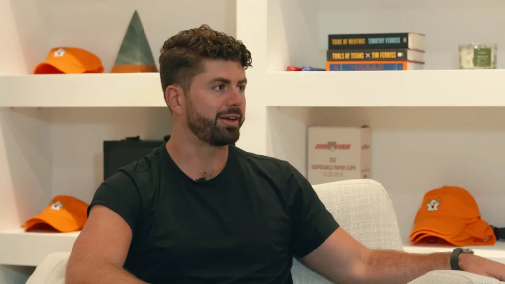 Bearded man with short, curly hair in black t-shirt sits on light-colored couch. White shelves with orange hats, books, and various items behind him. Man is contemplating how to make money writing. Cozy room setting ideal for creative thoughts and productivity. Creatorpoint – Creator Knowledge, Simplified. The best resources, podcast summaries, and learning for creators.