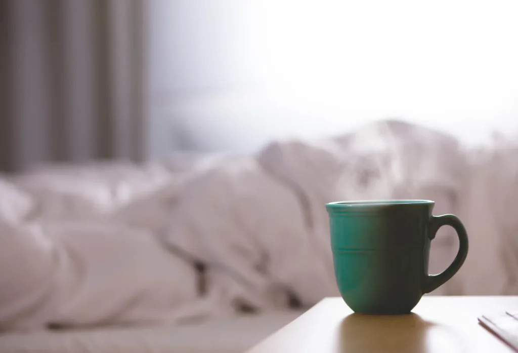 Green ceramic mug on a wooden table, white rumpled bed in the background suggesting a relaxed morning setting ideal for contemplating how to make money writing; soft natural light from the left. Creatorpoint – Creator Knowledge, Simplified. The best resources, podcast summaries, and learning for creators.