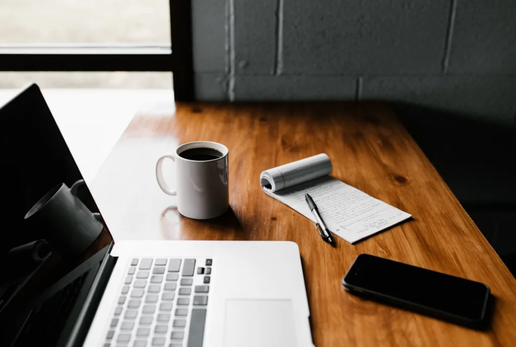 Alt Text: Inspiring home office workspace setup with natural light, featuring a wooden desk, laptop with tips on making money writing, white coffee mug, handwritten notepad, pen, and smartphone. Creatorpoint – Creator Knowledge, Simplified. The best resources, podcast summaries, and learning for creators.