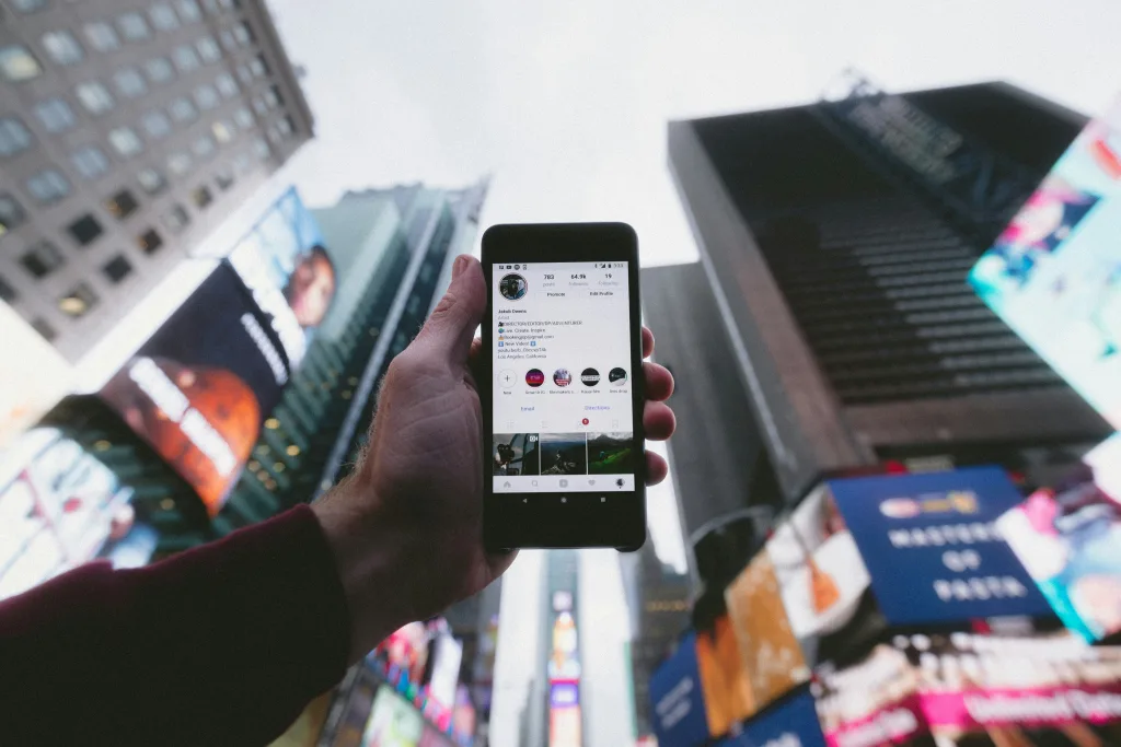 Person displaying Instagram profile on smartphone in urban city with tall buildings and billboards. Focus on phone screen showing multiple posts, account info, links to latest YouTube videos, podcast episodes on Creatorpoint. Ideal for social media influencers, digital content creators seeking exposure and engagement. Creatorpoint – Creator Knowledge, Simplified. The best resources, podcast summaries, and learning for creators.