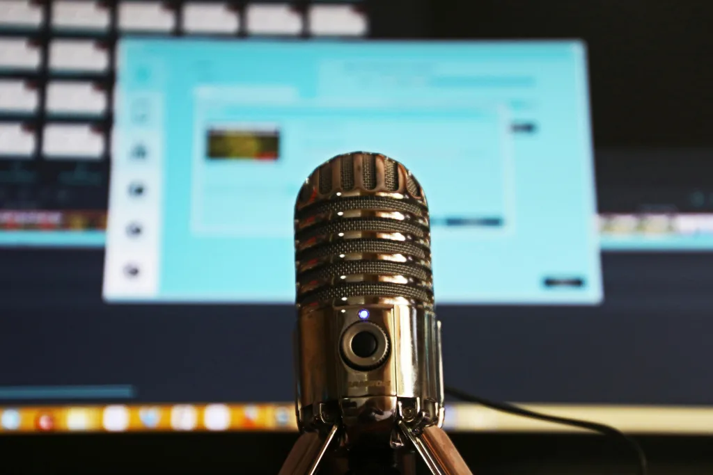 Alt text: "Close-up black podcast microphone with metallic accents on stand, positioned in front of a computer screen displaying blurred video or audio editing software, ideal for professional podcast creators seeking clarity. Creatorpoint – Creator Knowledge, Simplified. The best resources, podcast summaries, and learning for creators.