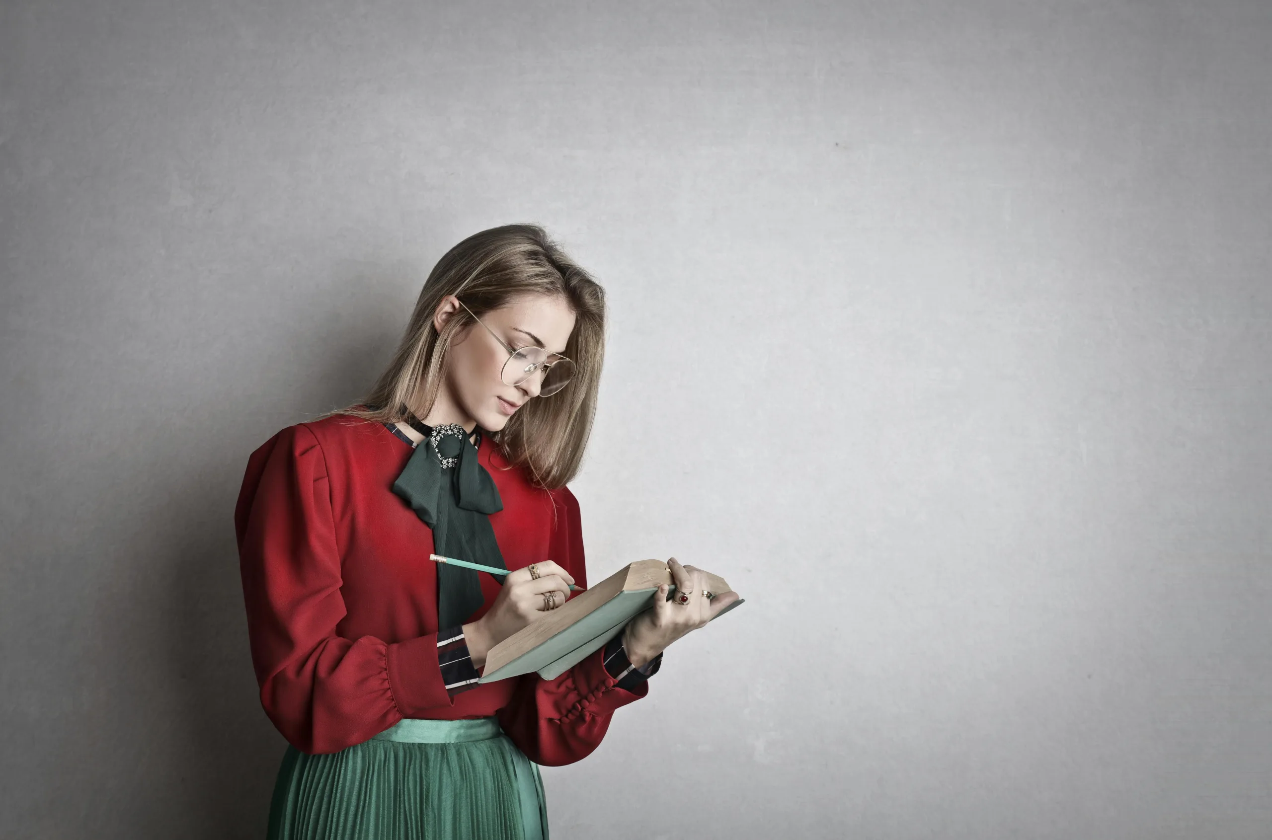 Alt text for Optimal Indexing: "Blonde woman with glasses in red blouse and green skirt writing ideas in notebook, gray background, content creator brainstorming or podcast summary preparation Creatorpoint – Creator Knowledge, Simplified. The best resources, podcast summaries, and learning for creators.