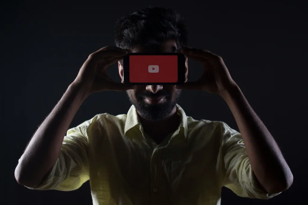 Person in light shirt holding smartphone with glowing YouTube logo on screen against dark background, symbolizing digital content creation entry point Creatorpoint – Creator Knowledge, Simplified. The best resources, podcast summaries, and learning for creators.