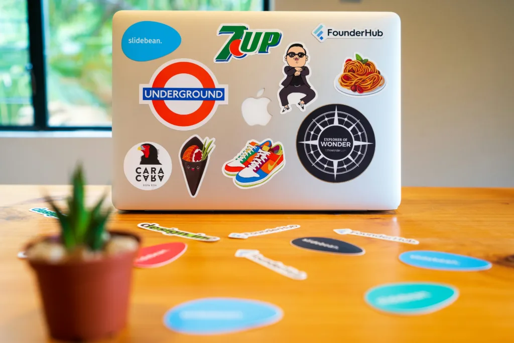Colorful laptop with 7Up logo, sunglasses illustration, and spaghetti plate sticker on a wooden table. Small potted plant in foreground. Perfect cozy creator space setup for podcast recording. Ideal home office inspiration featuring vibrant stickers, creative vibes, and inviting aesthetics. Creatorpoint – Creator Knowledge, Simplified. The best resources, podcast summaries, and learning for creators.
