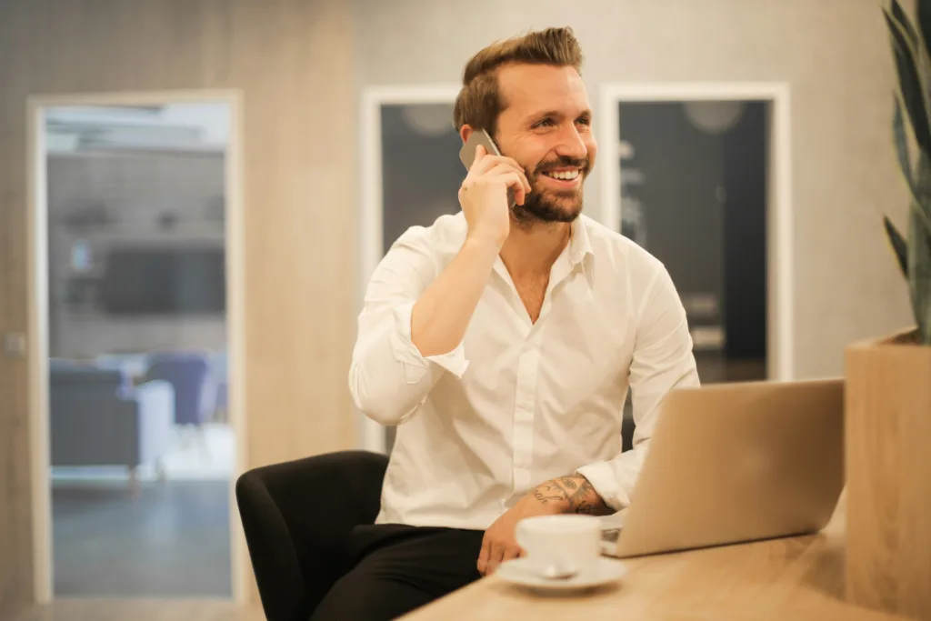 Entrepreneur in white shirt at desk, smiling while discussing solo business ideas on smartphone; open laptop beside coffee cup. Blurred modern office space with glass doors in background, conveying innovative and professional workspace environment. Ideal for articles on entrepreneurship and digital business trends. Creatorpoint – Creator Knowledge, Simplified. The best resources, podcast summaries, and learning for creators.