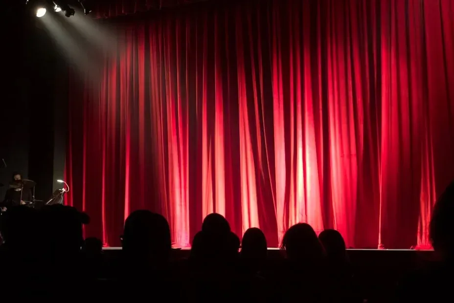 Dimly lit stage with closed red velvet curtains, silhouetted audience in foreground, spotlight from upper left corner on curtains. Anticipating upcoming performance or event, much like awaiting valuable YouTube tips and tricks to be revealed. Creatorpoint – Creator Knowledge, Simplified. The best resources, podcast summaries, and learning for creators.