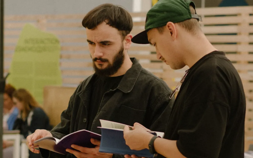 Two individuals reading books on starting a successful YouTube channel; one bearded in a black jacket, the other wearing a green cap and black shirt. Indoor setting focused study scene. Enhance your YouTube journey with expert advice and insights. Creatorpoint – Creator Knowledge, Simplified. The best resources, podcast summaries, and learning for creators.