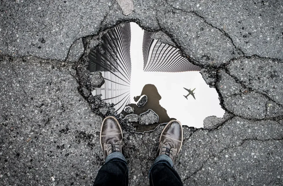 Urban perspective: stylish brown shoes beside water-filled pothole reflecting city skyscrapers and airplane overhead; silhouette on cracked pavement evokes travel vlog vibe. Creatorpoint – Creator Knowledge, Simplified. The best resources, podcast summaries, and learning for creators.