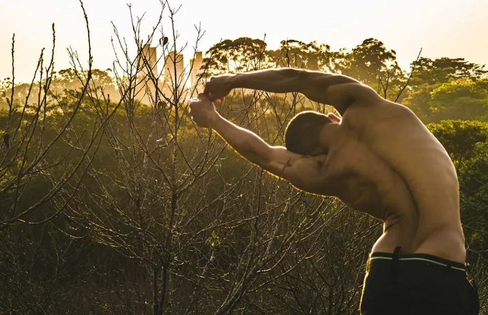 Shirtless person stretches outdoors with arms interlocked above head, embodying freedom and focus for solo business inspiration. Positioned before bare trees, sunlight filters through dense foliage in background. Perfect representation of creativity and clarity away from distractions. Creatorpoint – Creator Knowledge, Simplified. The best resources, podcast summaries, and learning for creators.