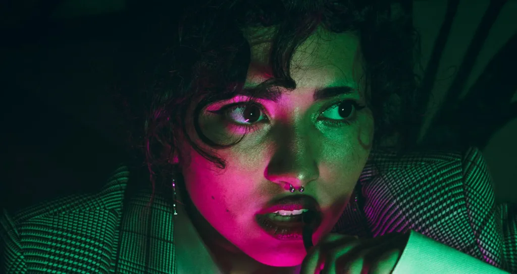 Thoughtful person with curly hair and nose ring illuminated by green and pink lighting, holding a pen near mouth, pondering storytelling improvement techniques. Dramatic portrait showcasing deep reflection and creativity. Creatorpoint – Creator Knowledge, Simplified. The best resources, podcast summaries, and learning for creators.
