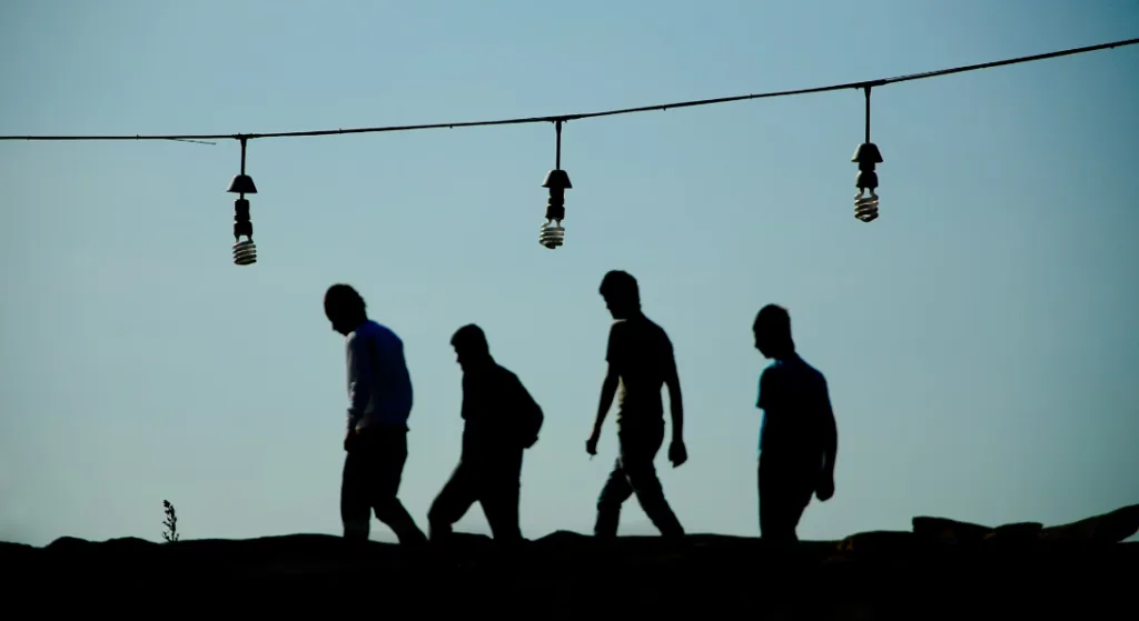 Silhouettes of four figures walking on a ridge under a clear blue sky symbolize the journey to becoming a better storyteller. Hanging lightbulbs illuminate their path, representing progression and creativity as they move right. Embrace storytelling skills and creative growth in this illustrative image. Creatorpoint – Creator Knowledge, Simplified. The best resources, podcast summaries, and learning for creators.