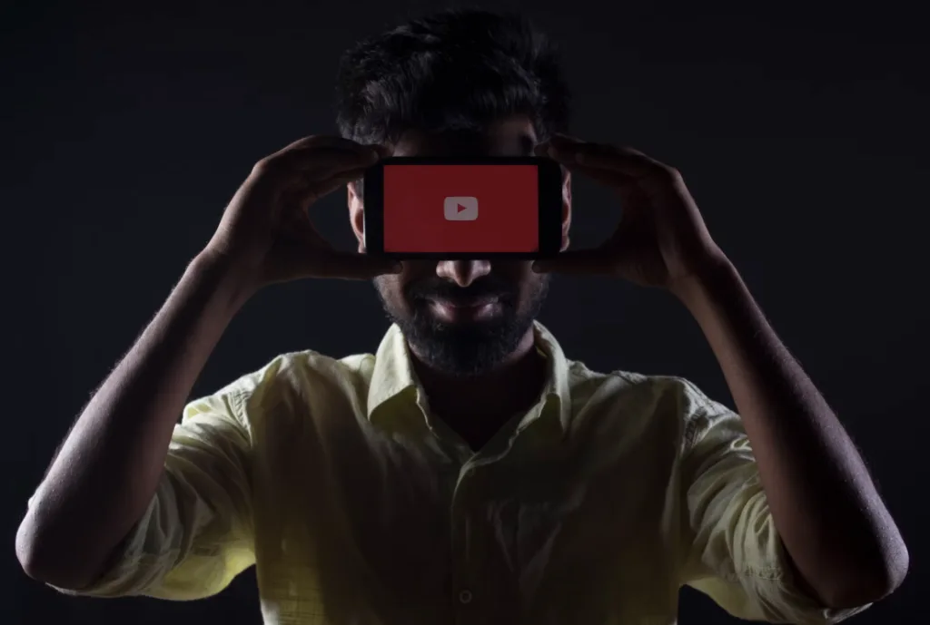 A person with a light-colored shirt holds a smartphone horizontally, obscuring their face. The phone displays the YouTube logo on a red screen, suggesting they might share YouTube tips and tricks. The background is dark. Perfect for tutorials or educational content related to YouTube strategies. Creatorpoint – Creator Knowledge, Simplified. The best resources, podcast summaries, and learning for creators.