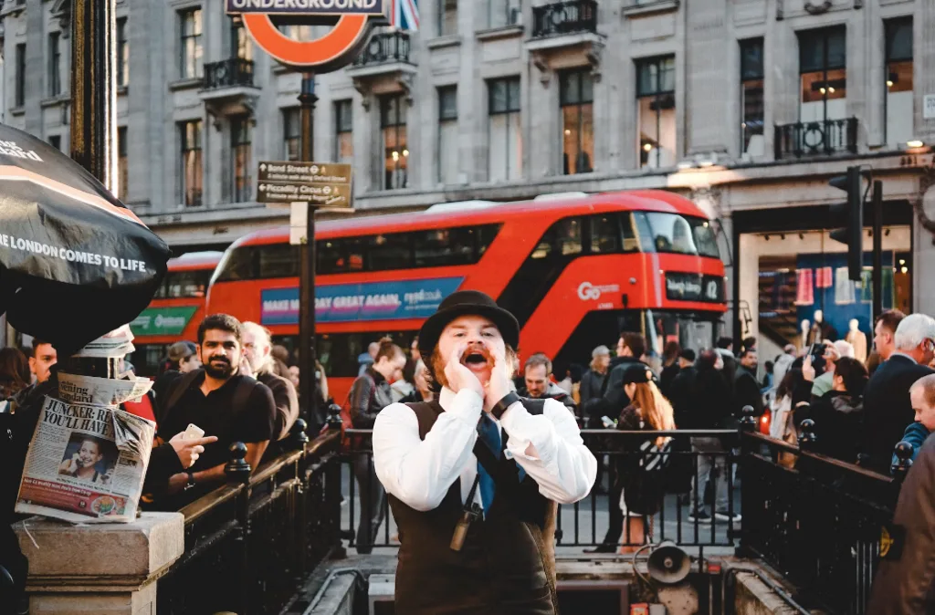 Victorian-clad person dramatically shouts near a London Underground entrance as a red double-decker bus passes. Faceless YouTube channel ideas buzz in the crowd's chatter. Newspapers on a stand complete the lively scene, capturing London's vibrant street life and digital creativity. Creatorpoint – Creator Knowledge, Simplified. The best resources, podcast summaries, and learning for creators.