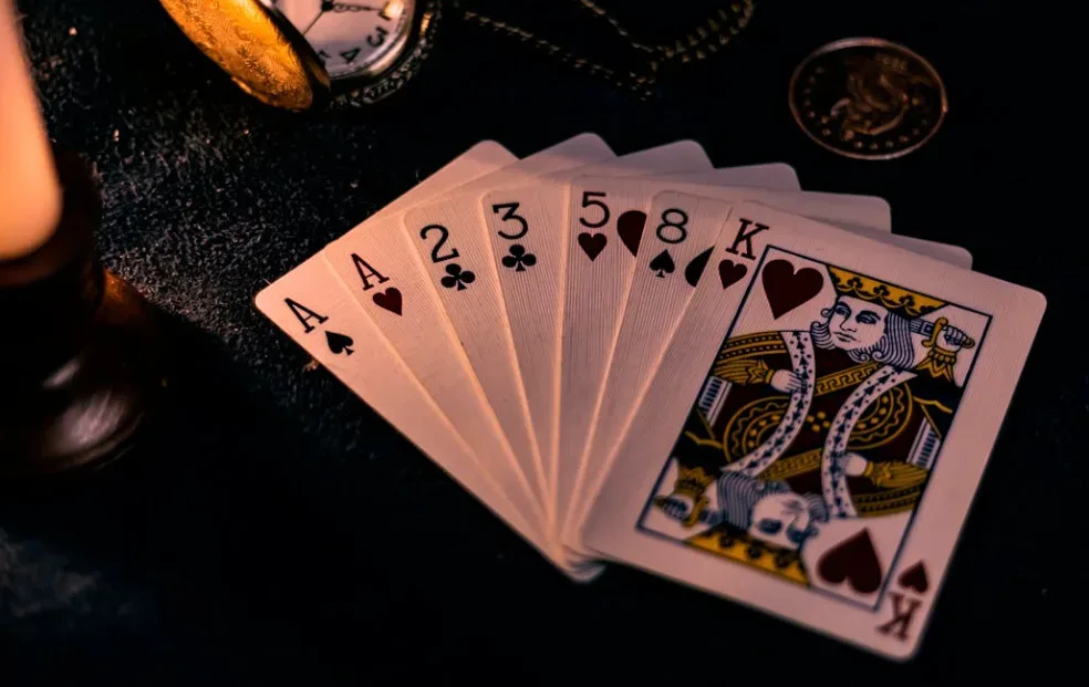 Alt text: A hand displays two aces of hearts, two of spades, three of clubs, five of diamonds, eight of spades, king of hearts; nearby are a pocket watch and coin on a dark surface. Ideal for card magic enthusiasts. Perfect faceless YouTube channel idea for magic content in poker or card tricks. Creatorpoint – Creator Knowledge, Simplified. The best resources, podcast summaries, and learning for creators.