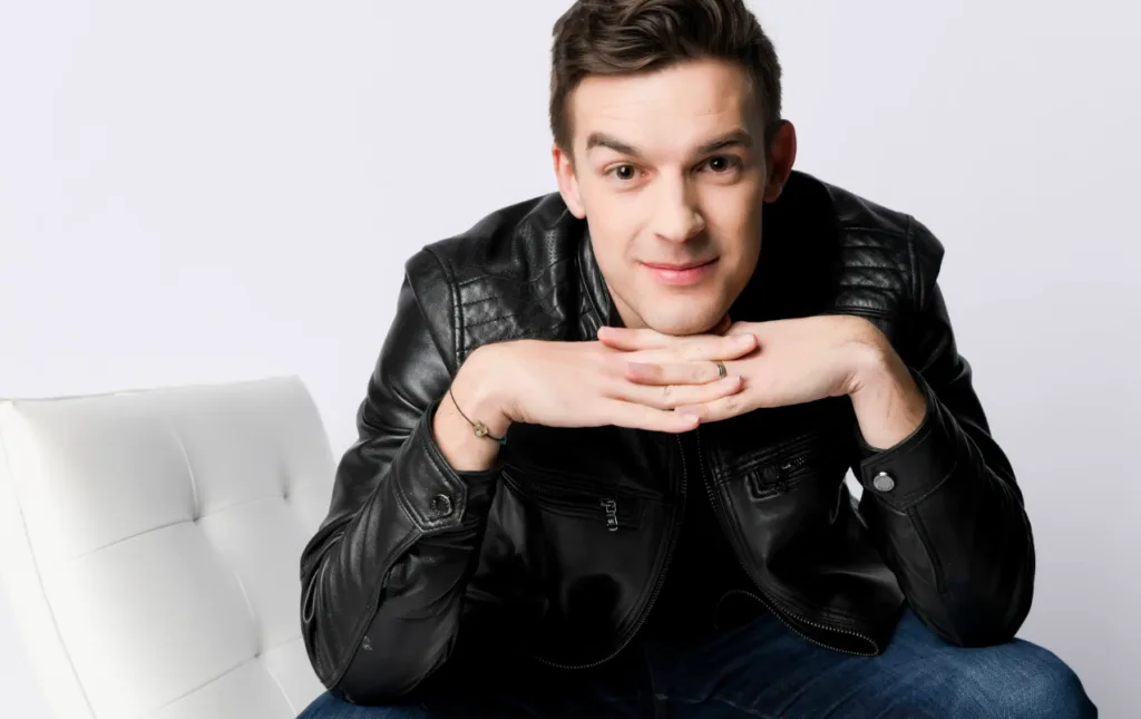 Person in black leather jacket and blue jeans sitting on white chair, resting chin on hands, short brown hair, neutral expression. They ponder strategies for gaining views on YouTube Shorts. Plain white background enhances focus on individual's thoughtful gaze directly at camera. Creatorpoint – Creator Knowledge, Simplified. The best resources, podcast summaries, and learning for creators.