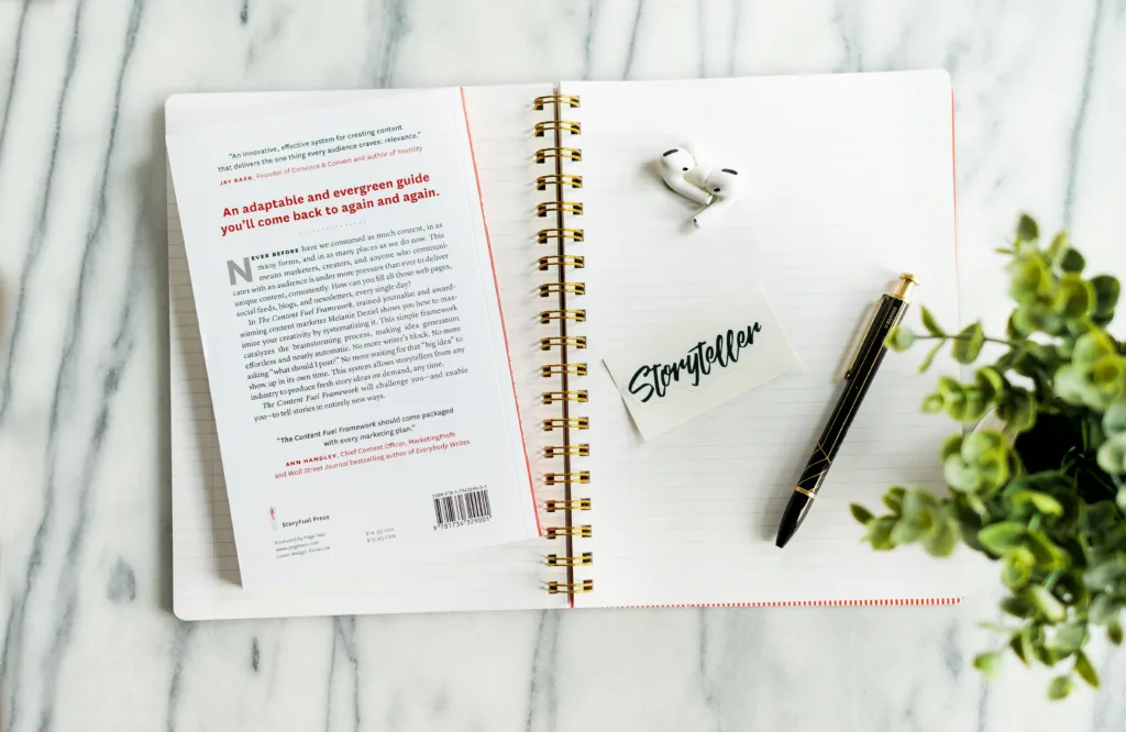 Open spiral notebook on marble surface featuring a book, wireless earbuds, pen, and card titled "How to Be a Better Storyteller" on the right page; small plant visible in corner. Perfect setup for storytelling enthusiasts seeking inspiration and creativity boost. Creatorpoint – Creator Knowledge, Simplified. The best resources, podcast summaries, and learning for creators.