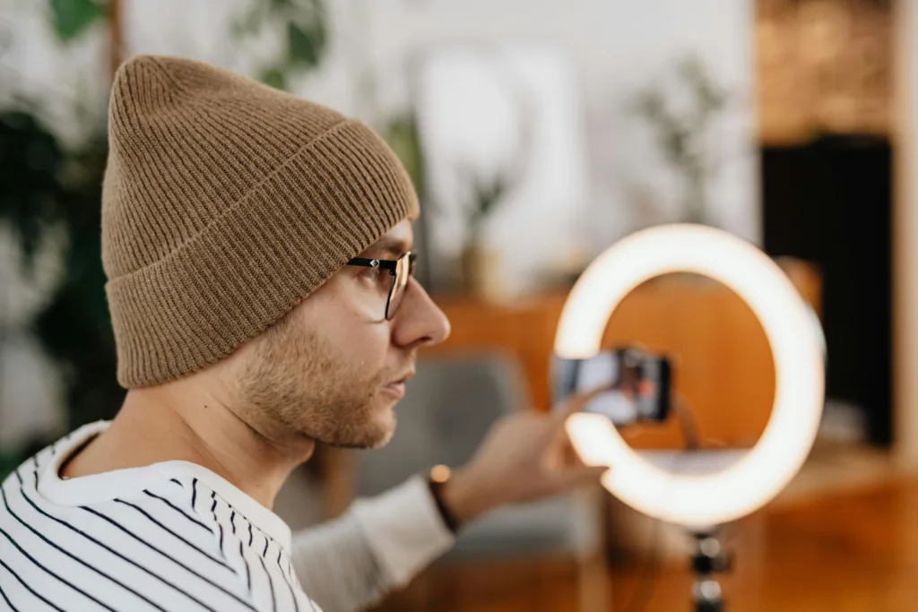 Person wearing brown knit hat and glasses indoors, focused on learning YouTube Shorts optimization on a smartphone mounted to a tripod with ring light; blurry indoor plants and decor in background. Creatorpoint – Creator Knowledge, Simplified. The best resources, podcast summaries, and learning for creators.